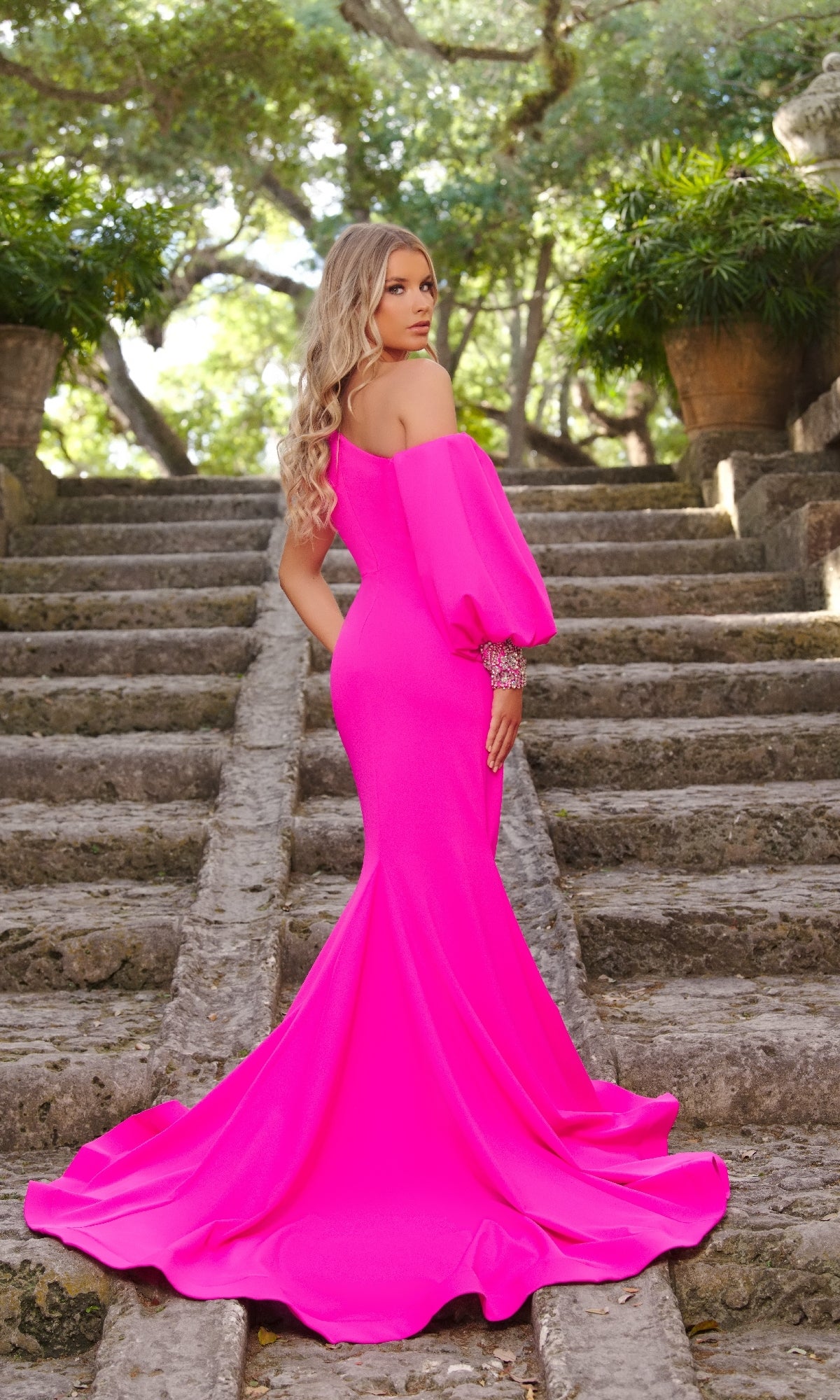 A woman in the Ava Presley 38352 one-sleeve long prom dress stands on stone steps outdoors, surrounded by greenery and large potted plants.